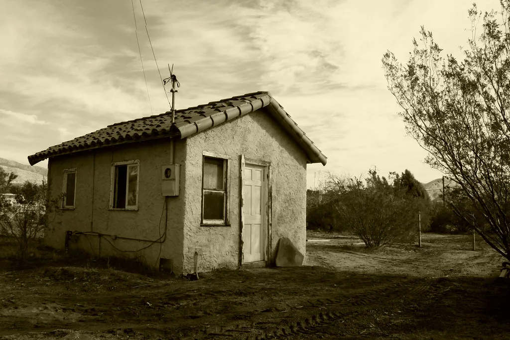 Small pale building with empty windows on dry ground, in sepia.