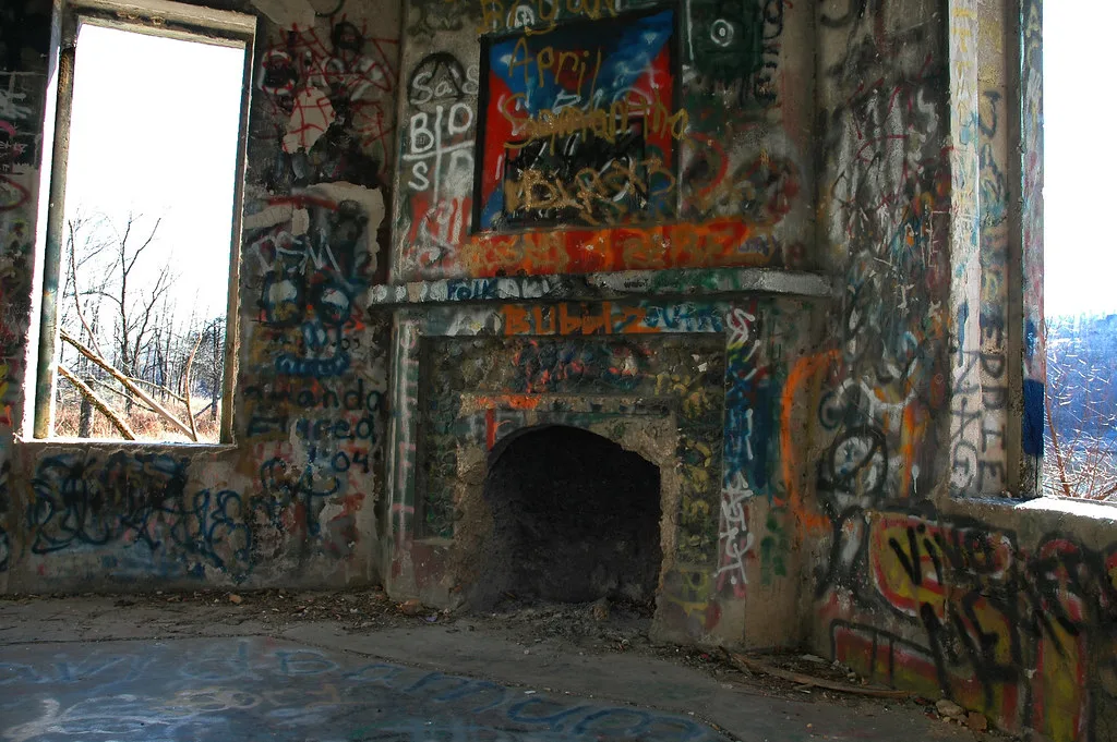 Cheminée d’une pièce abandonnée couverte de graffitis entre deux grandes fenêtres vides ouvertes sur les arbres.