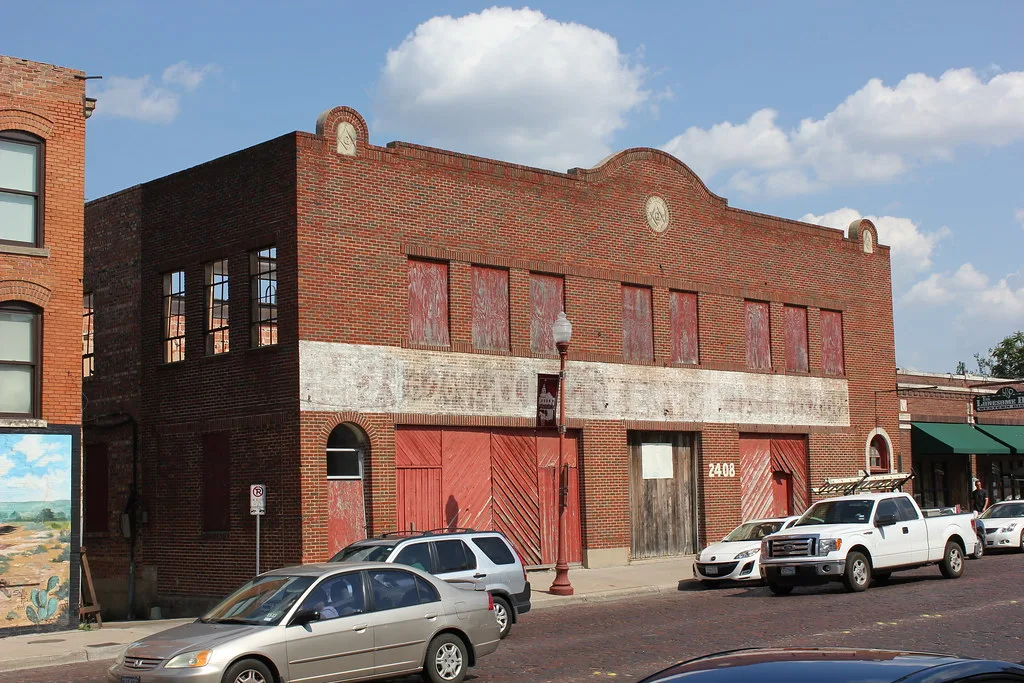 Old brick building with boarded openings behind parked cars.