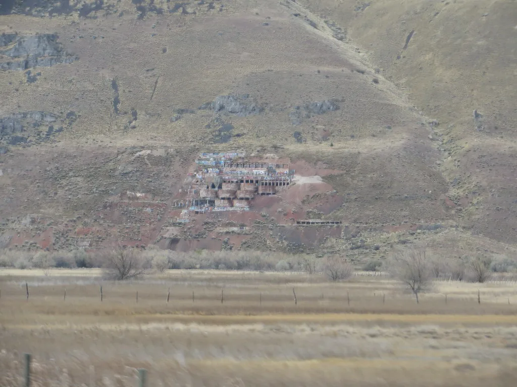 Abandoned industrial ruins with empty openings and graffiti on an arid hillside beyond a meadow.
