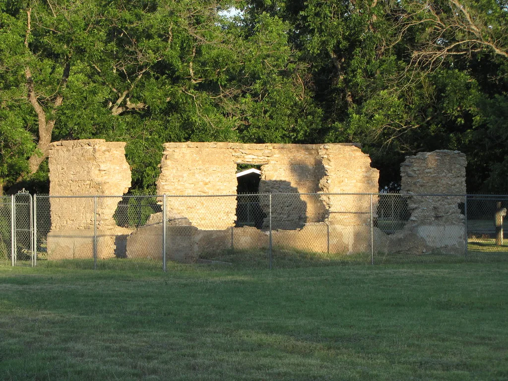 Roofless ruins of a small stone building behind a fence, lit by low sunlight.
