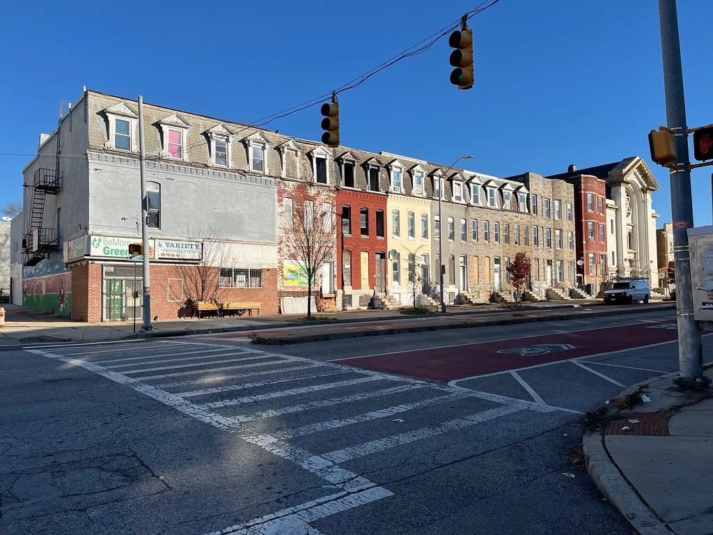 Urban row of abandoned houses with boarded or empty openings and deteriorated dormers, facing a sunny intersection.