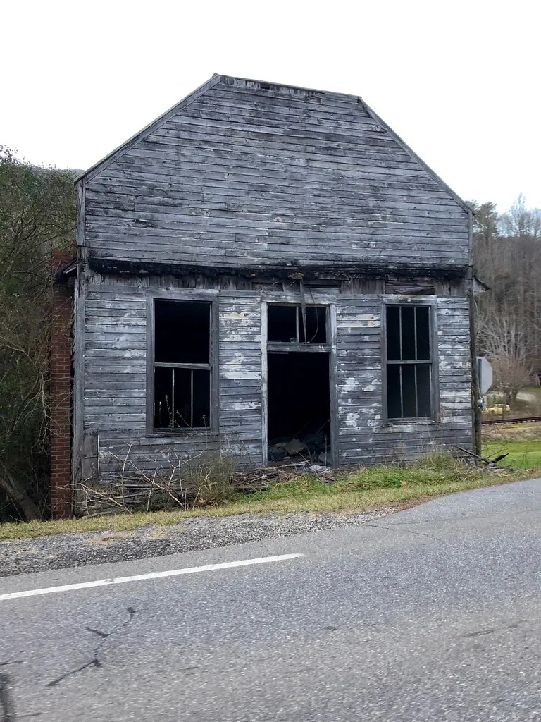 Old gray wooden shop with empty openings beside a road.