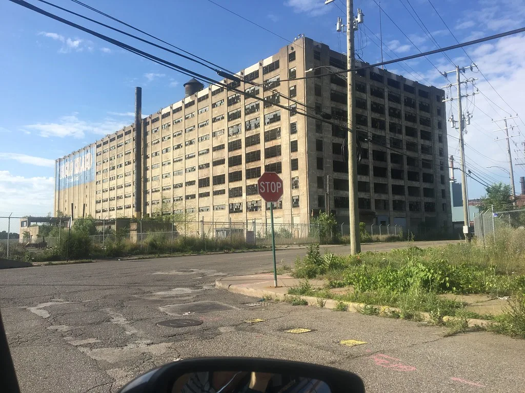 Grande usine aux fenêtres brisées derrière une clôture et des herbes hautes.