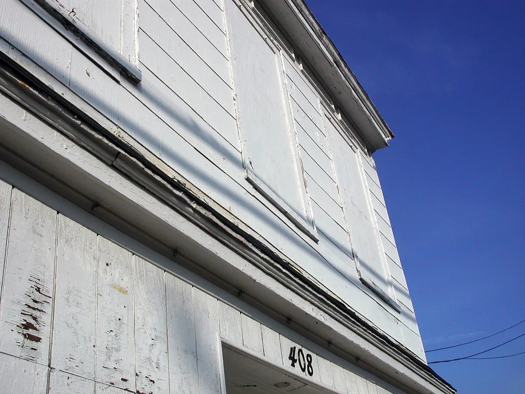 White wooden facade with boarded windows and a deteriorated cornice.