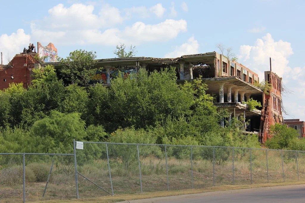 Ruins of a large brick and concrete building overgrown with trees behind a fence.