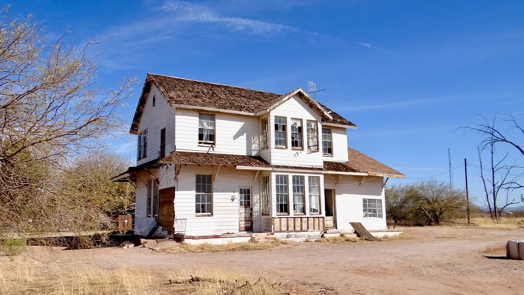 White two-storey abandoned house with broken windows and a deteriorated porch in a dry landscape.