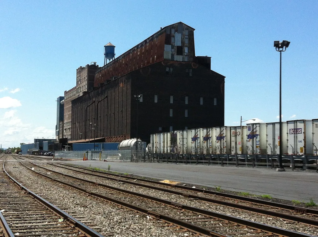 Large disused industrial building with sealed openings and damaged cladding beside railway tracks.