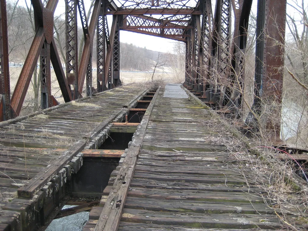 Rusty former steel railway bridge with decayed wooden ties and large holes in the deck.