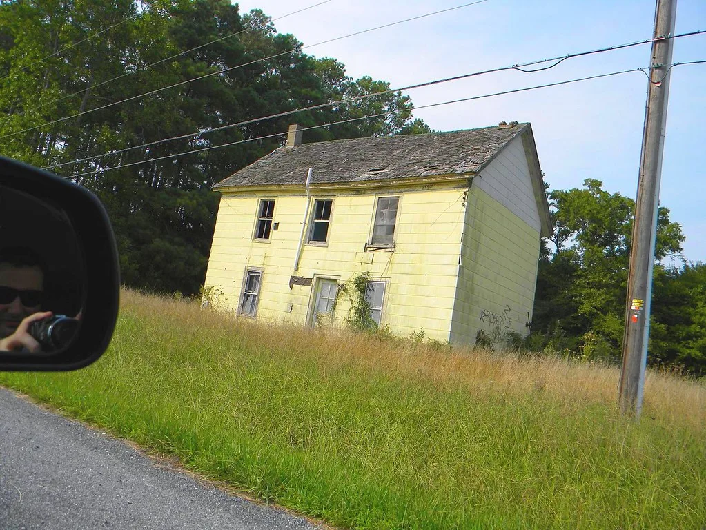 Abandoned yellow house with broken window panes and a damaged roof in tall grass beside a road.
