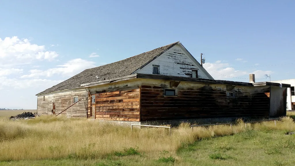 Long neglected wooden building with a peeling white gable and brown extension in a meadow.