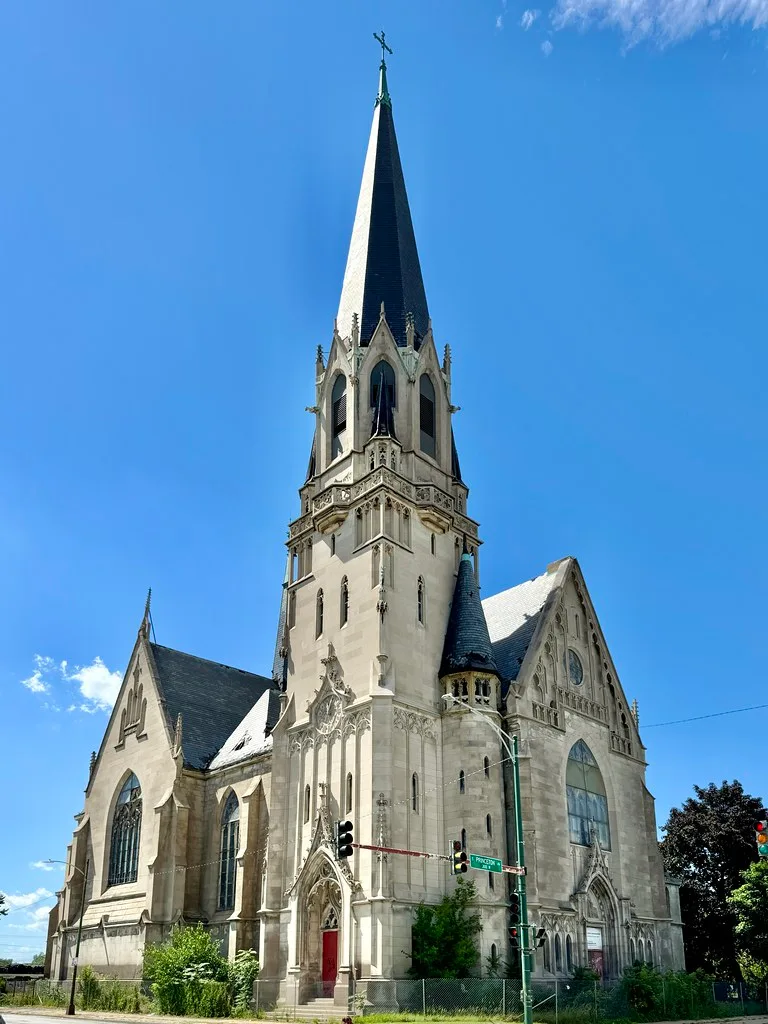 Old church with a tall spire, boarded entrances and vegetation at its base.