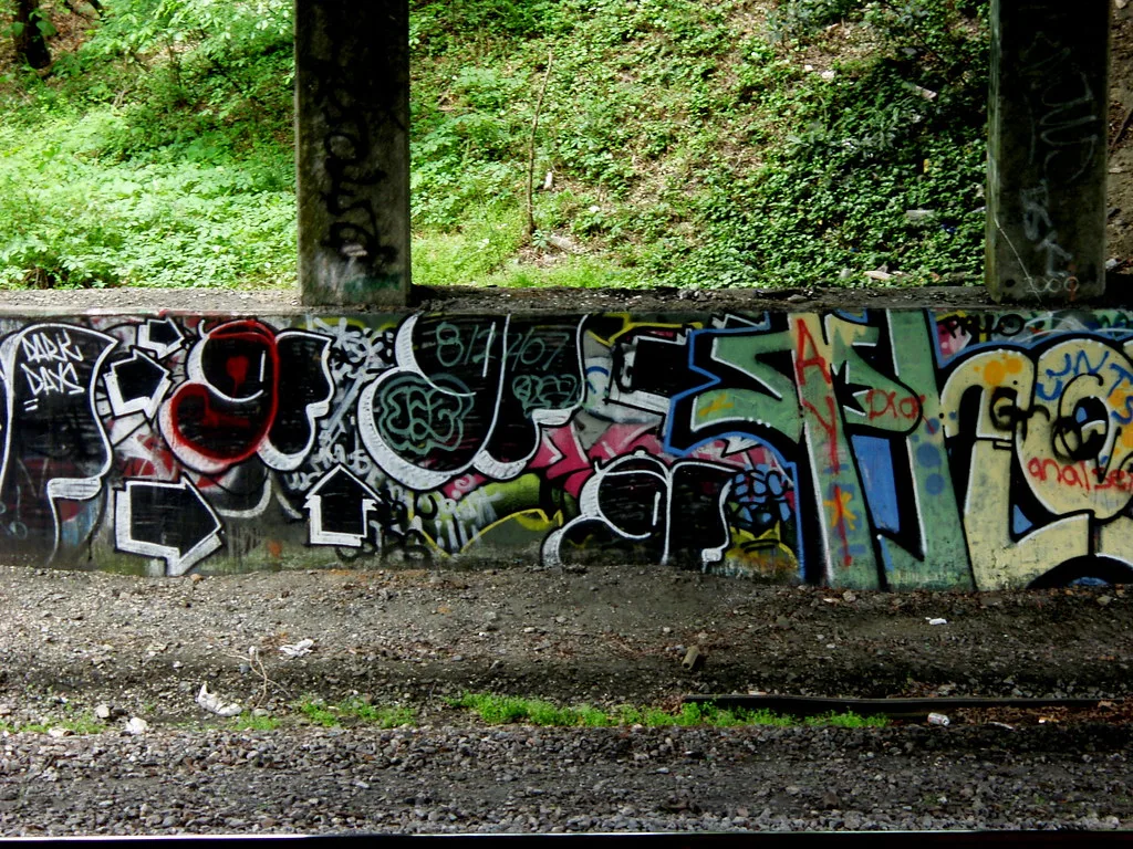 Piliers et muret de béton couverts de graffitis près d’un talus végétalisé.