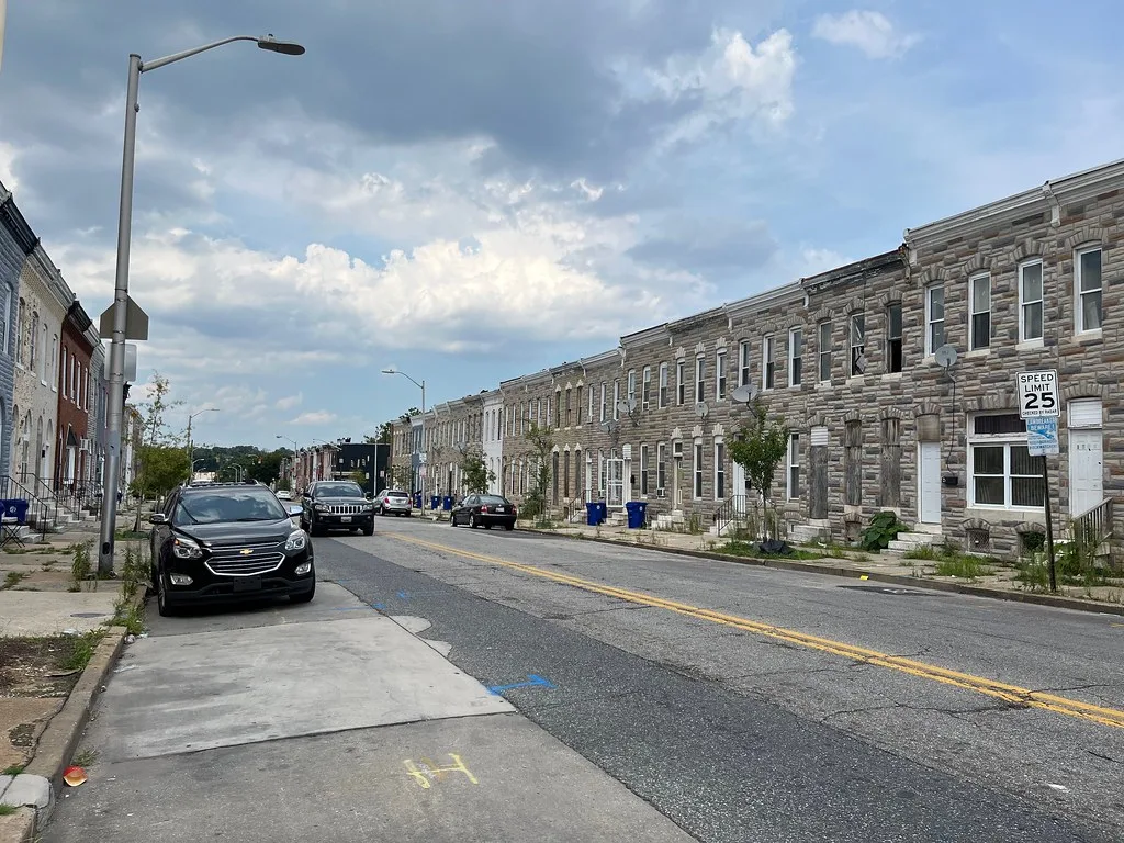 Street lined with houses, several with boarded windows and weed-covered entrances.