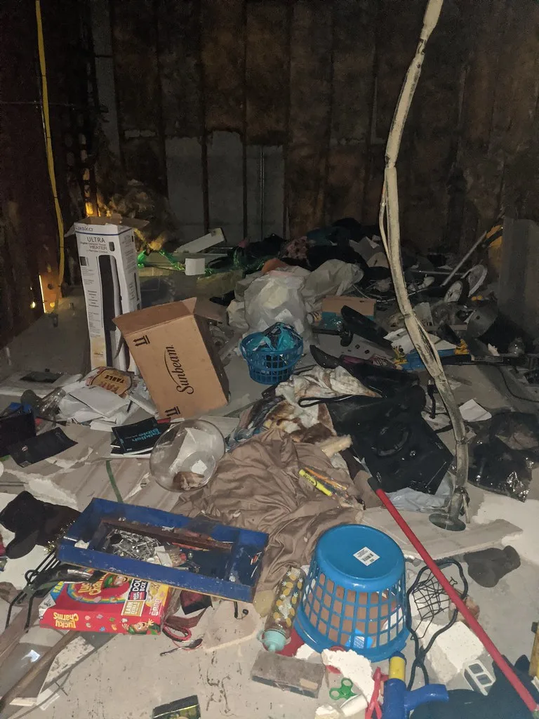 Pièce abandonnée aux parois nues et sol couvert de cartons, textiles et débris sous une gaine pendante.