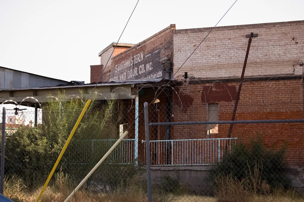 Former brick workshop with boarded windows and an overgrown covered loading platform behind a fence.