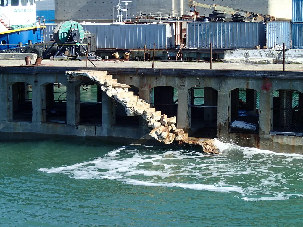 Broken concrete staircase descending to flooded openings in a ruined harbour structure.