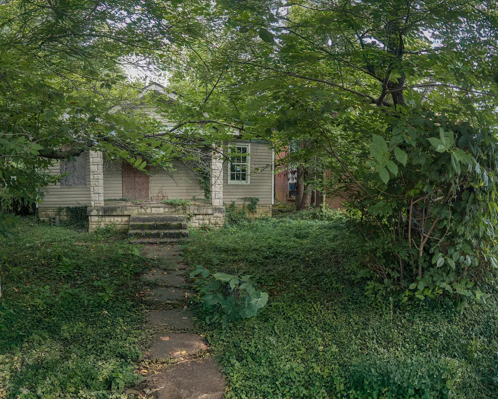 Old house with boarded openings behind dense vegetation.