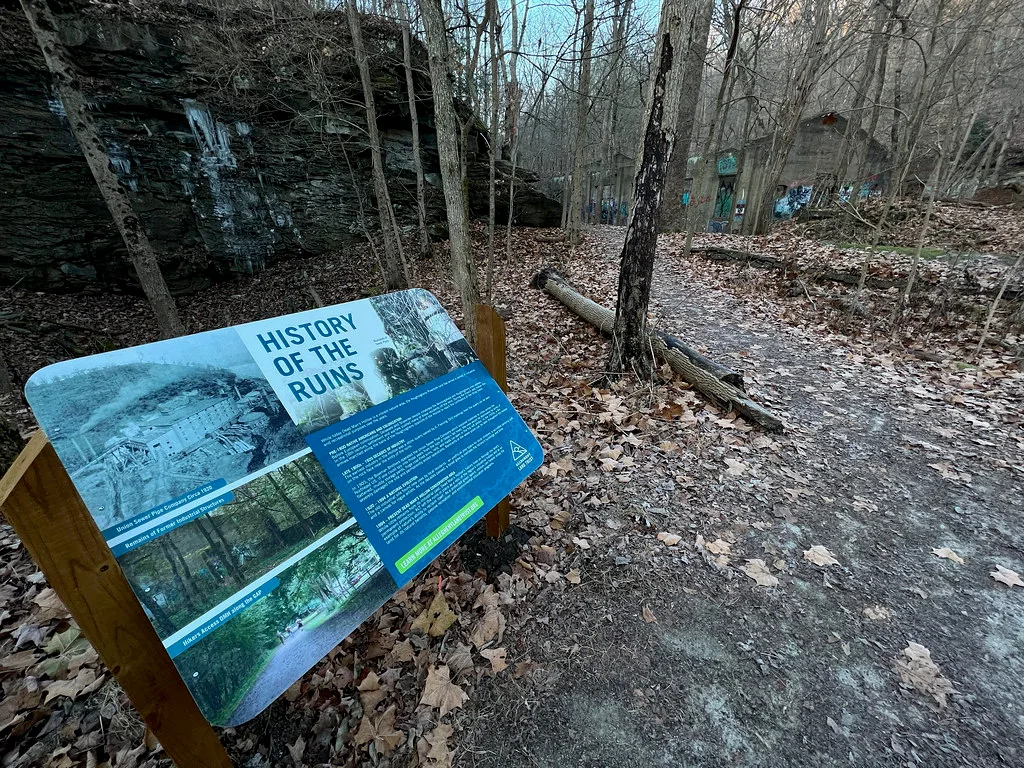 Ruines en forêt derrière un panneau explicatif au bord d’un sentier.