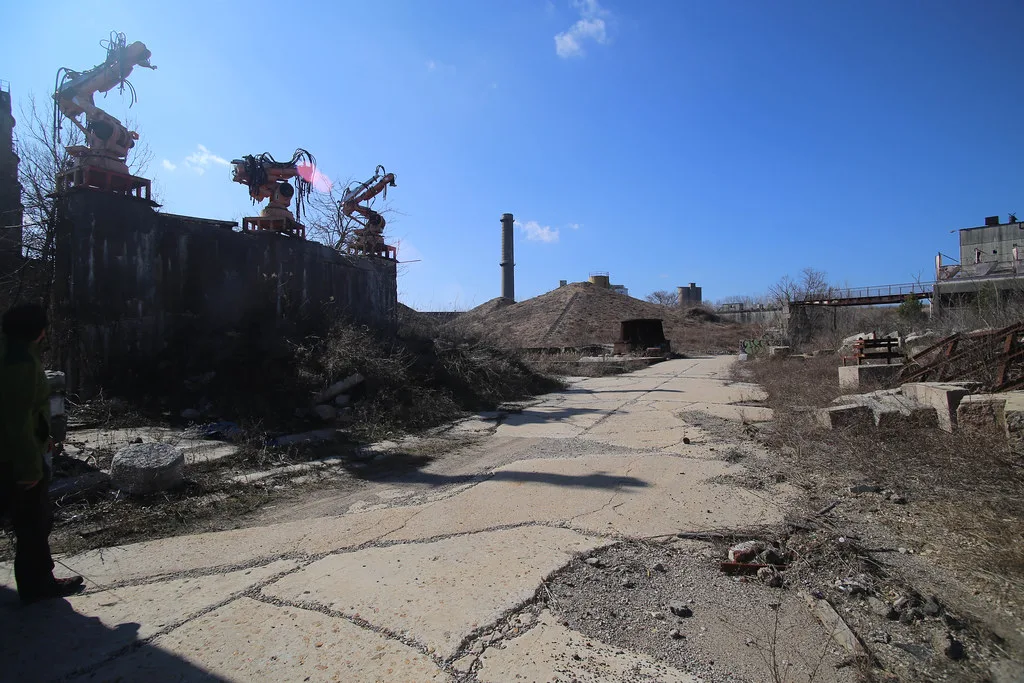 Derelict industrial site with rusty cranes, chimneys and a cracked concrete lane overgrown with vegetation.