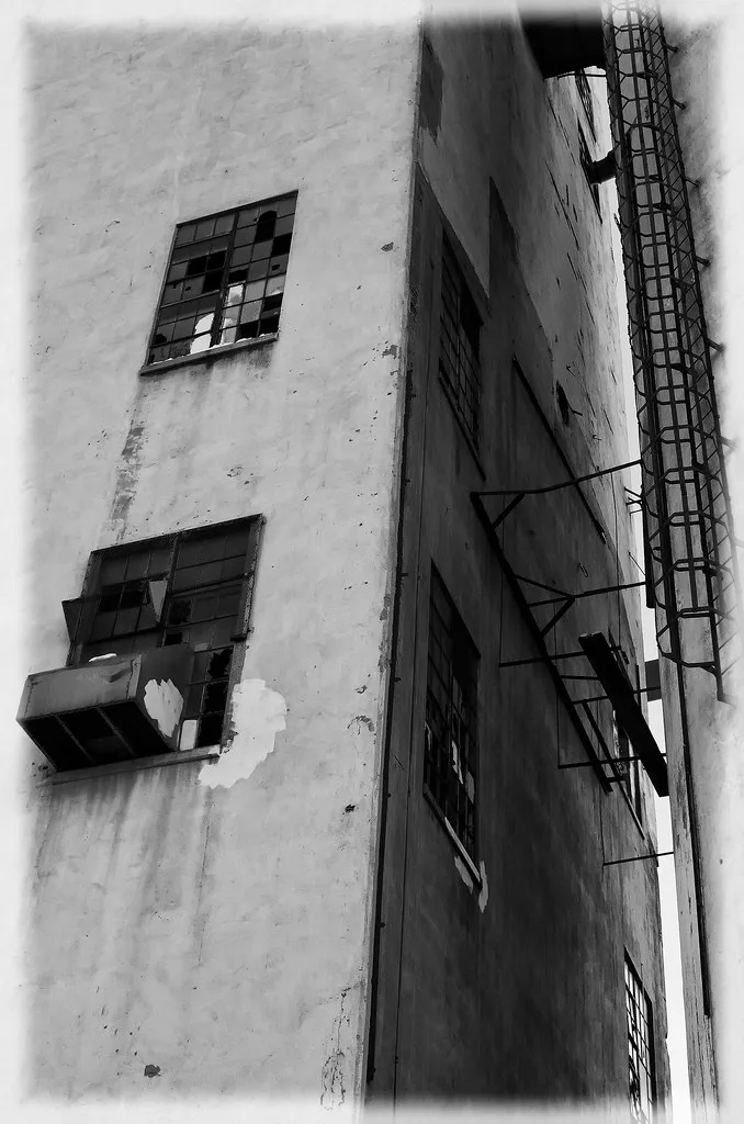 Industrial facade with broken windows and an exterior ladder, in black and white.