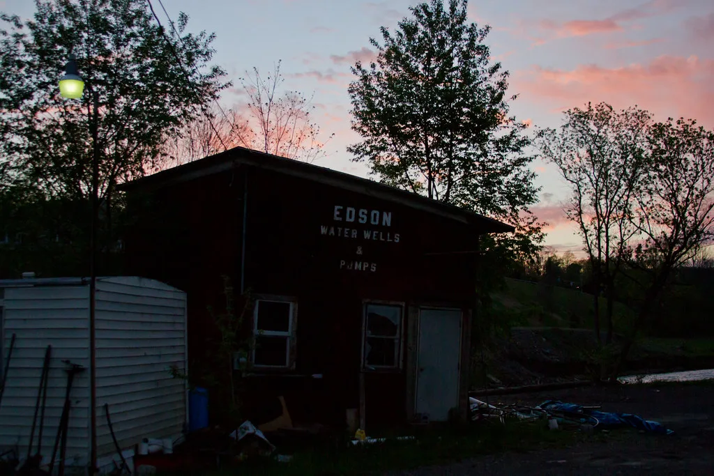 Petit atelier abandonné de bois sombre aux fenêtres brisées, ancienne inscription de pompes et débris devant la façade au crépuscule.
