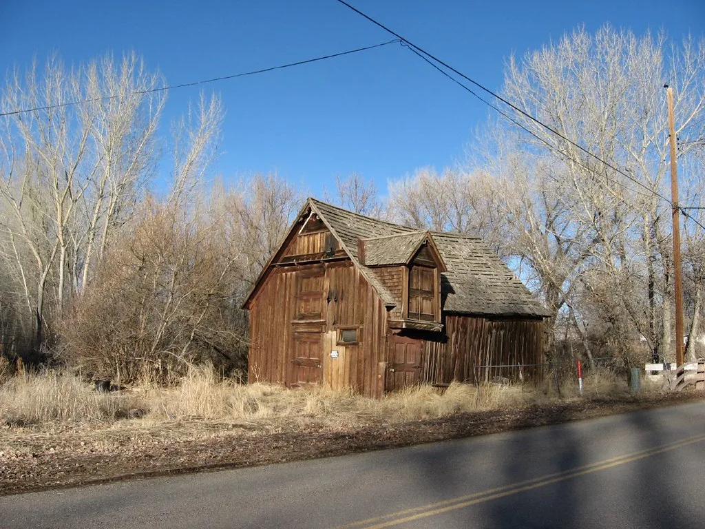 Small abandoned brown wooden building with a dormer, a damaged roof and closed doors amid dry grass.