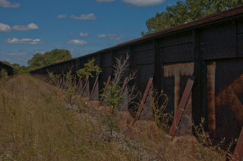 Former industrial passage between long rusty riveted steel girders, overgrown with tall grass and shrubs.