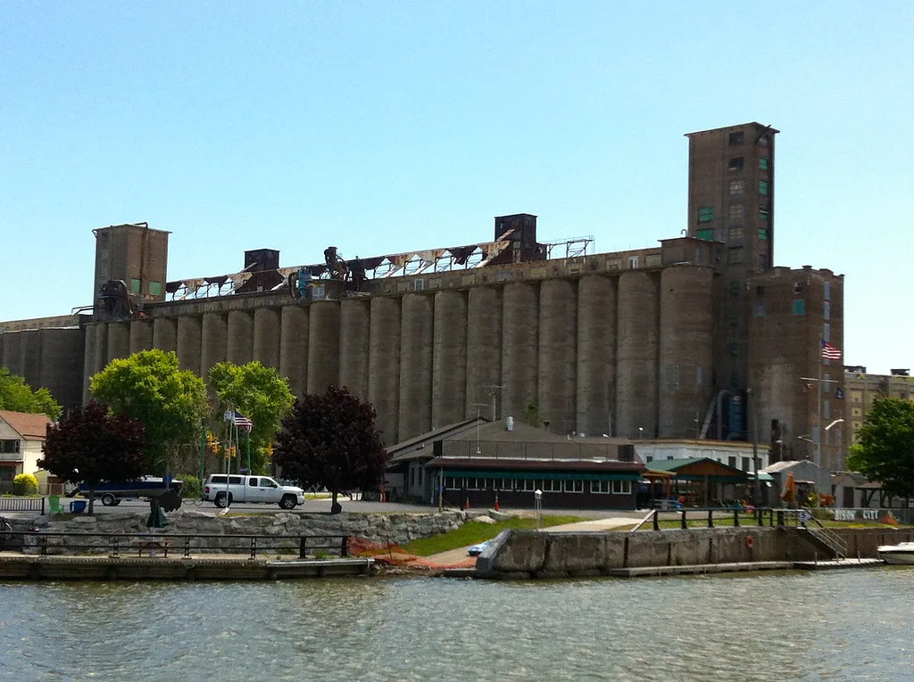Old concrete silos with damaged upper equipment beside the water.