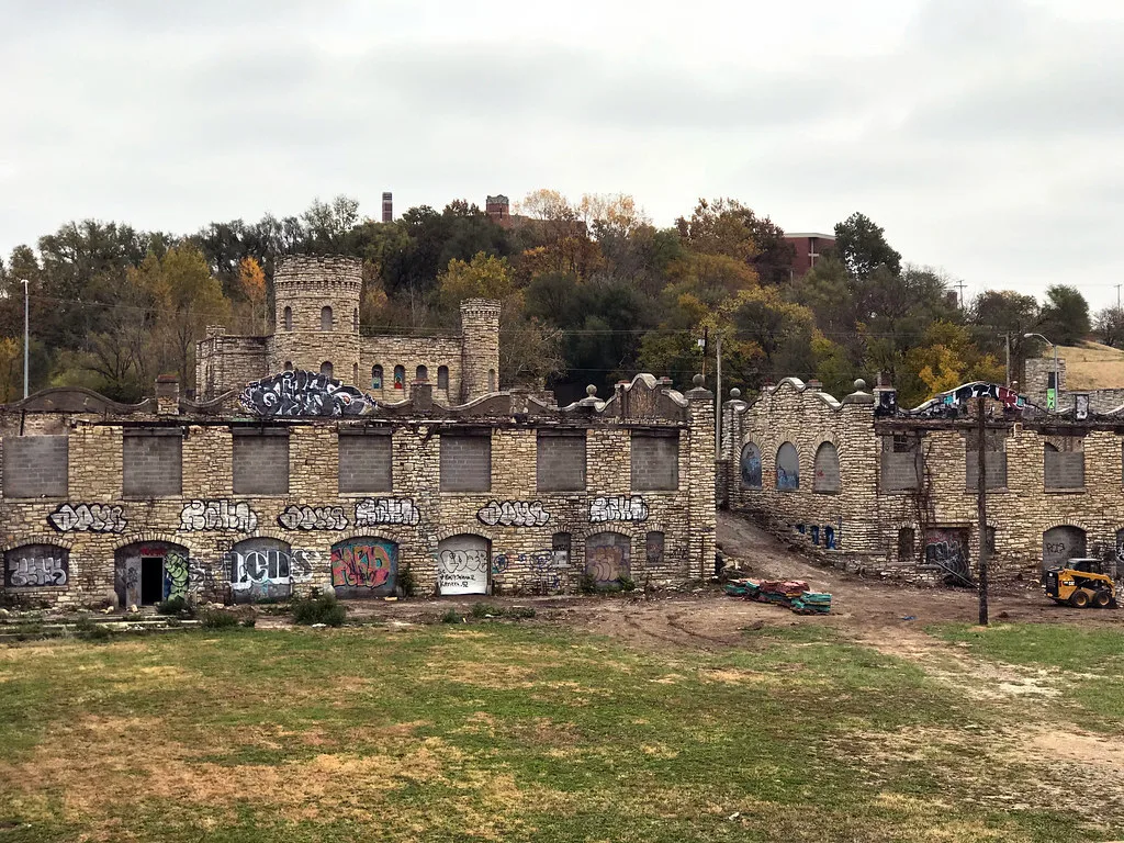 Stone ruins of a large building with towers and graffitied roofless facades before a wooded hill.