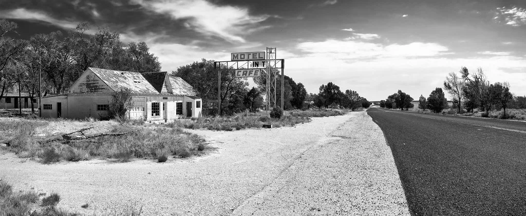 Abandoned former motel and cafe beside an empty road beneath a decayed metal sign, in black and white.