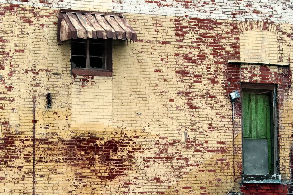 Brick facade with peeling yellow paint and a damaged green door.