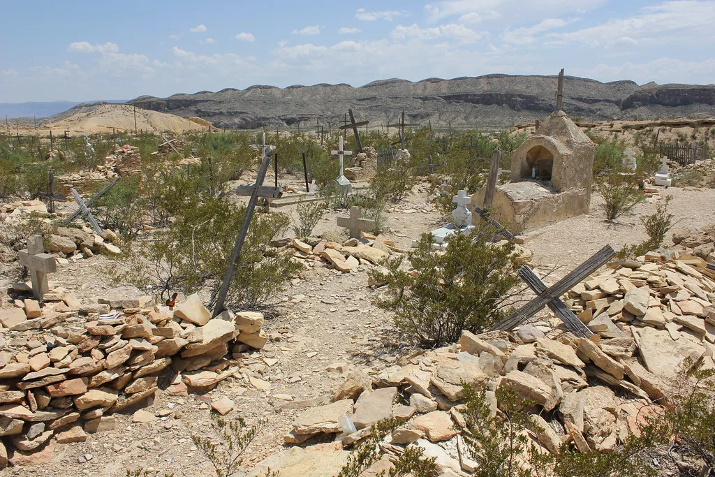 Broken stone graves and leaning wooden crosses among scrub in a desert cemetery.