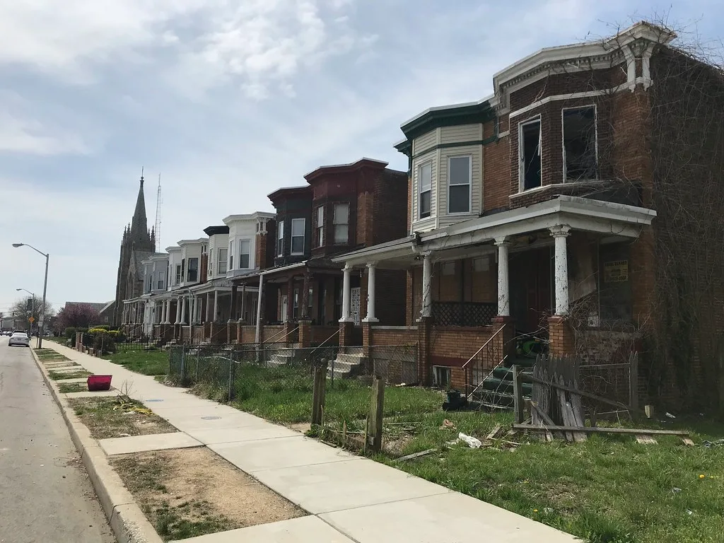 Maison abandonnée de briques à bow-window vide et porche dégradé au bout d’une rangée urbaine.