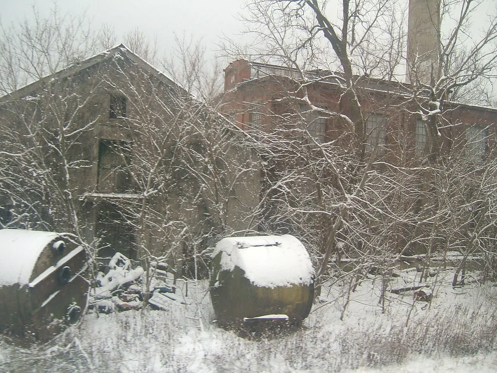 Abandoned brick industrial buildings among snow-covered trees, with two old tanks in the foreground.