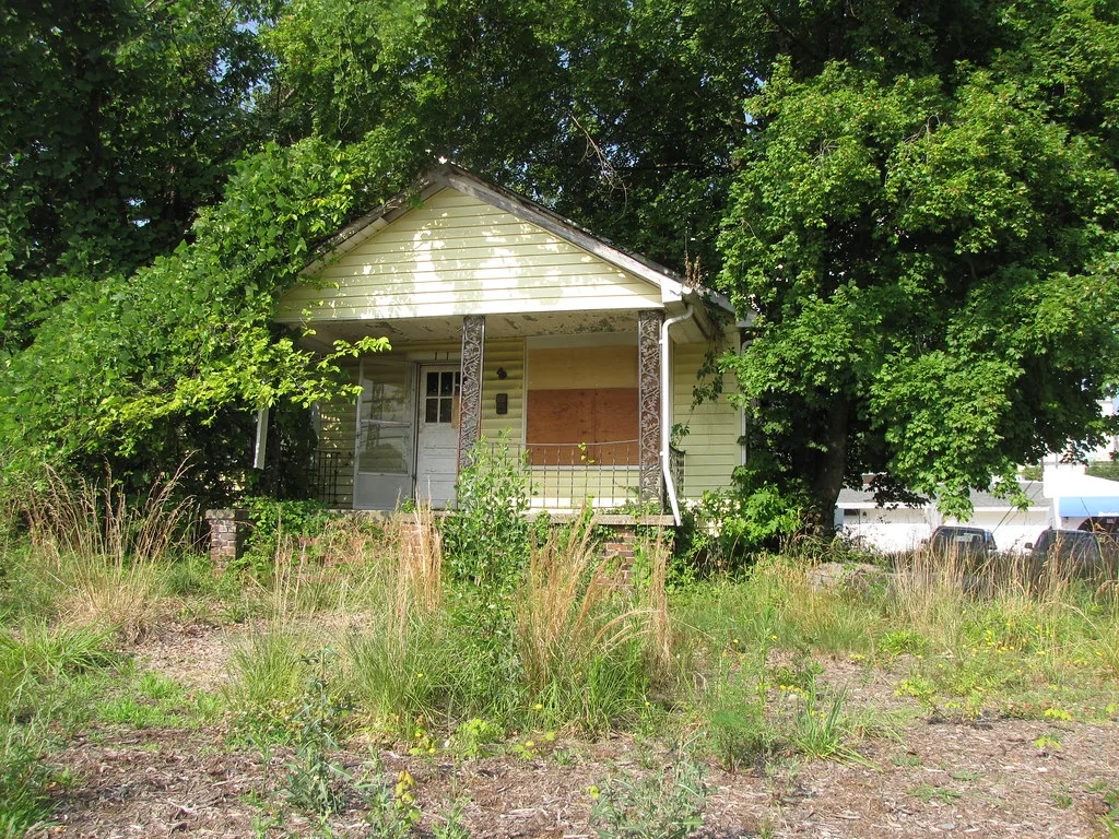 Petite maison jaune abandonnée à fenêtre condamnée et porche envahi de végétation sous les arbres.