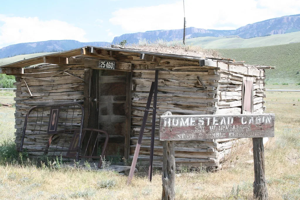 Abandoned rough wooden cabin with separated wall boards and a partially missing roof, with a bed frame leaning against the front.