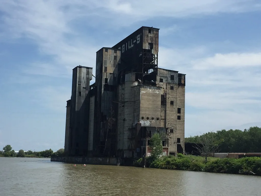 Grands silos de béton dégradés au bord de l’eau avec structures métalliques mises à nu.