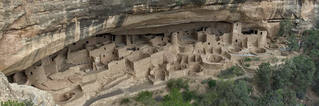 Ruins of stone buildings beneath a large overhanging cliff.