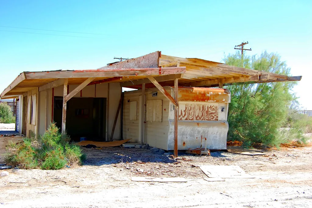 Small abandoned building with open walls beneath a deteriorated wooden canopy, with ground debris and shrubs on dry land.