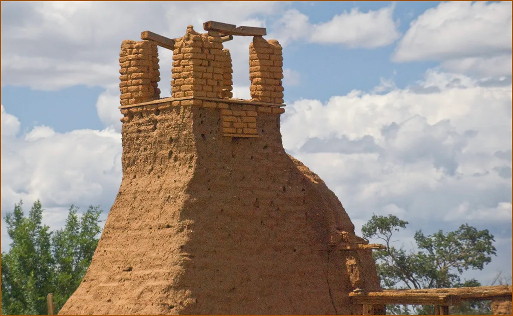Ruine en terre crue avec piliers de briques d’adobe et poutres apparentes sous un ciel nuageux.
