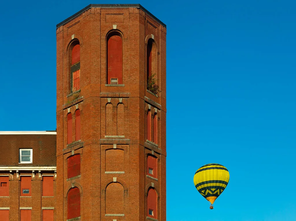 Abandoned brick tower with tall openings sealed by red panels and a hot-air balloon in the sky.