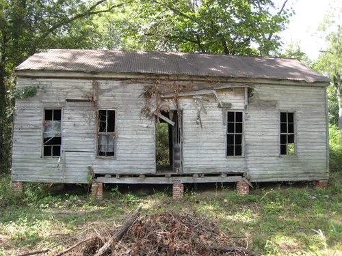 Small raised white house with broken windows and a missing door beneath trees.