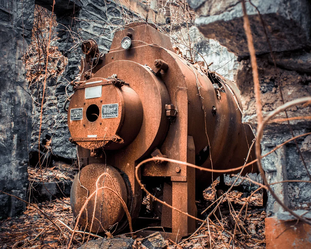 Ancienne chaudière rouillée entre des murs de pierre ouverts et des lianes sèches.