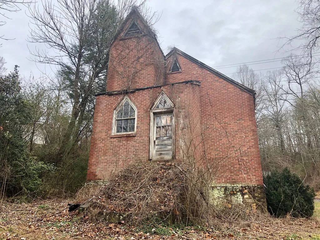 Small brick church with a decaying door overgrown with brambles.