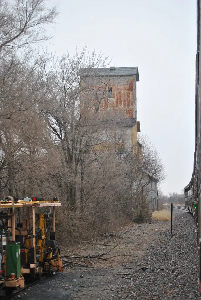 Ancien élévateur en bois partiellement caché par des arbres au bord des rails.