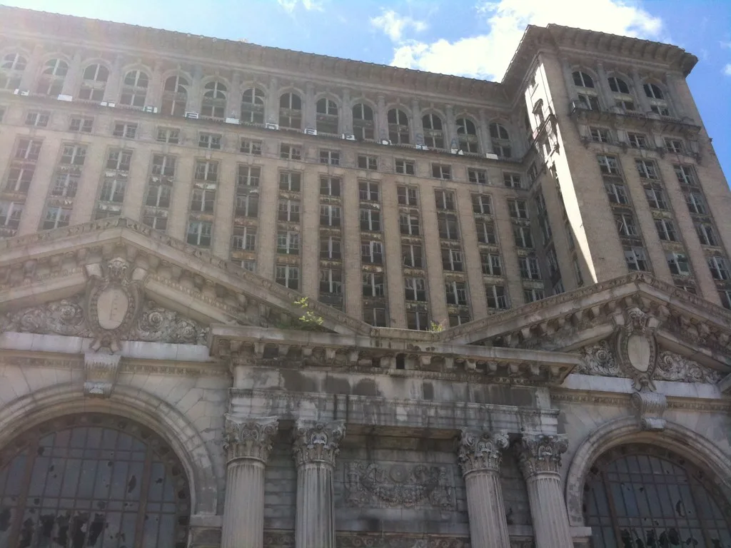 Grand bâtiment abandonné à façade monumentale de pierre, colonnes et fenêtres brisées avec des plantes sur les corniches.