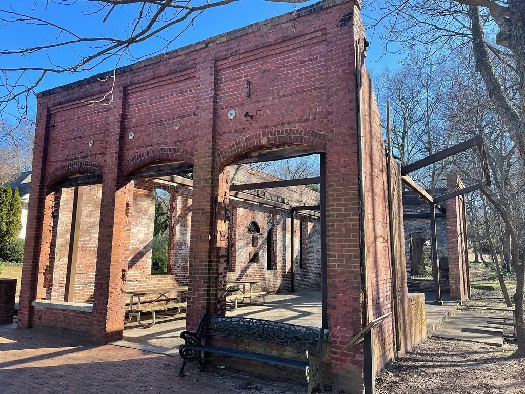 Preserved ruins of a roofless red brick building with large arched openings and exposed beams, with a bench and tables.
