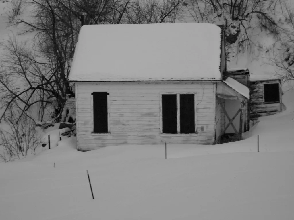 Small abandoned pale wooden house with empty windows and a deteriorated annex in the snow, in black and white.