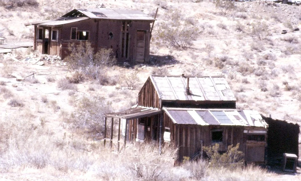 Deux cabanes abandonnées de bois aux fenêtres vides et toits de tôle abîmés sur une pente aride.